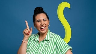 Ariana Baborie lachend mit Finger nach oben zeigend vor blauem Hintergrund „S“